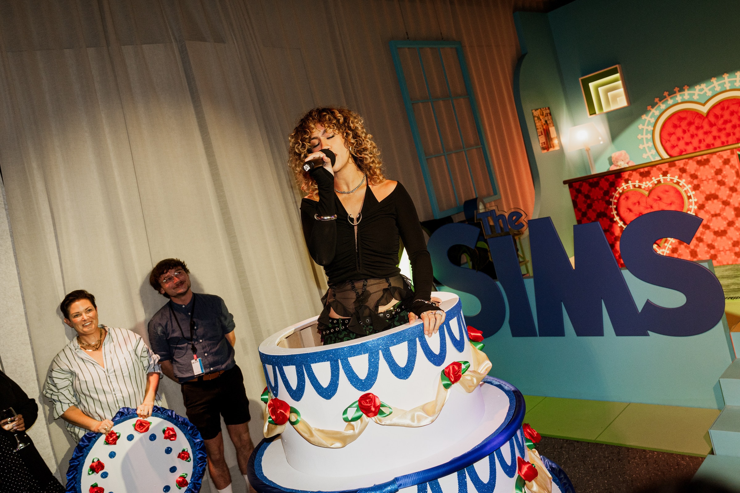 A performer bursting out of a giant birthday cake in front of The Sims logo
